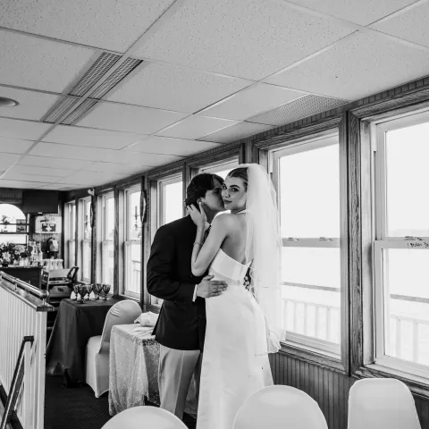 Bride and groom share intimate moment in a room with large windows.