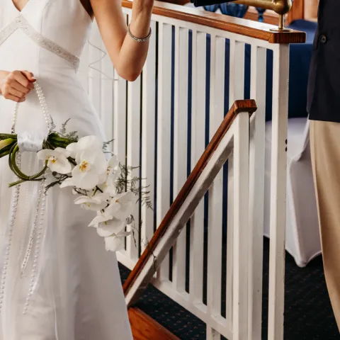 Bride in white dress next to suited person, holding hands, near a railing with flowers.