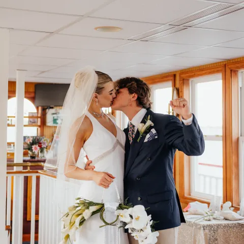 Bride and groom kissing indoors, she in white dress, he in navy blazer with flowers.