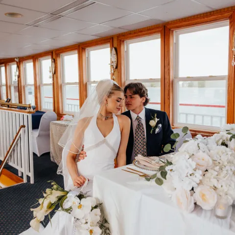 Bride and groom sitting at a table with flowers in a bright room.