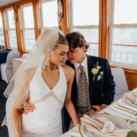 Bride and groom sitting closely at a dressed table on a boat with windows.
