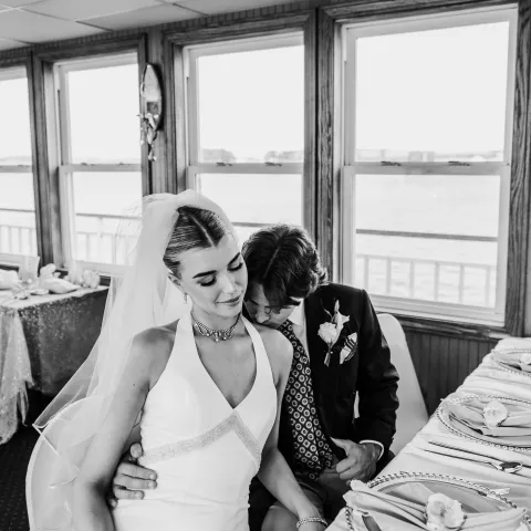 Bride and groom seated, holding hands in a room with windows overlooking water.