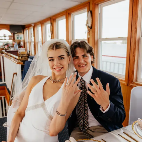 Bride and groom showing rings, seated indoors by windows.