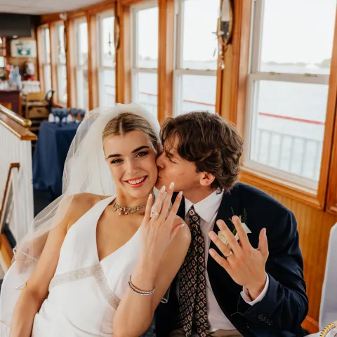 Bride and groom smiling, showing rings, groom kisses bride's cheek.