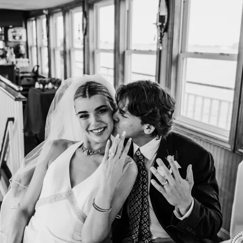 Bride and groom sitting, groom kisses bride's cheek, both show wedding rings.