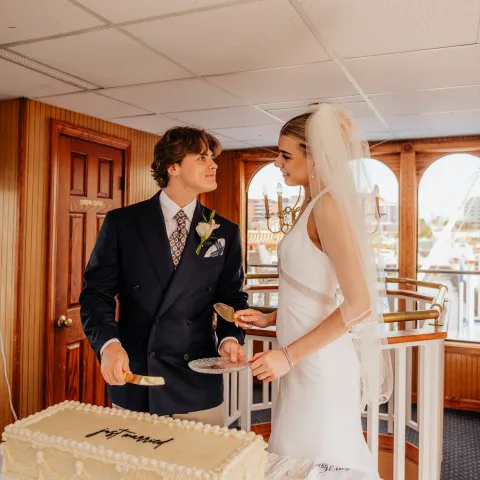 Couple cutting wedding cake on boat in elegant attire.