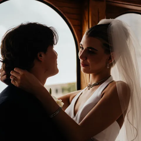 Bride and groom embrace, standing near large windows.