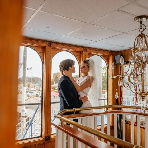Bride and groom embrace near large windows on a boat.