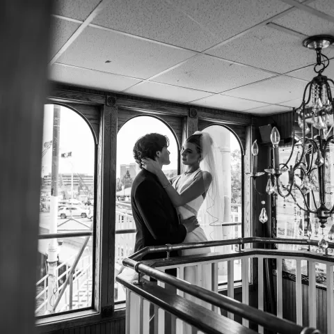Bride and groom embrace on indoor staircase with vintage chandelier and arched windows.