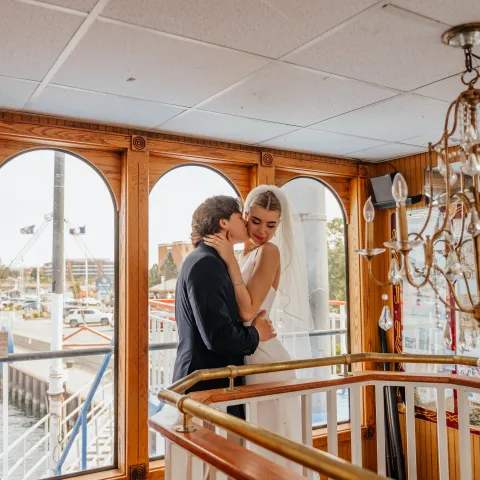Bride and groom embrace on a boat with large windows and a chandelier.