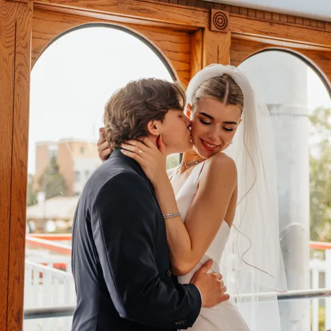 Bride and groom embracing by a window, groom kissing bride's cheek.