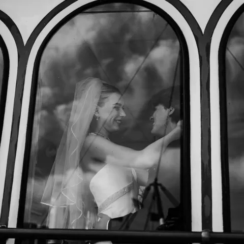Bride and groom smiling through arched window, with cloud reflections.