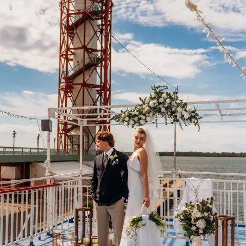 Couple in wedding attire on a boat deck with floral arch and tower background.