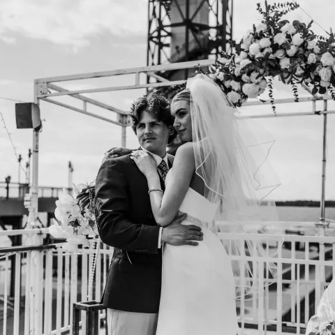 Bride and groom embracing under floral arch on outdoor deck.
