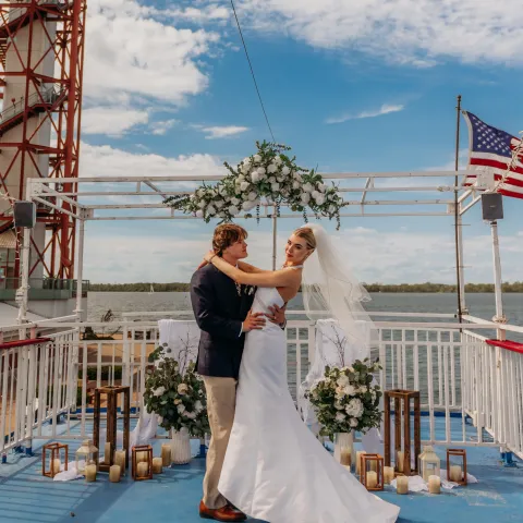 Bride and groom embrace on a boat deck with lighthouse and American flag in the background.