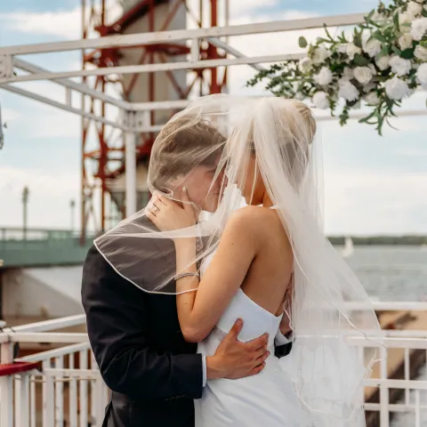 Bride and groom embrace under a veil near a waterfront and tower.
