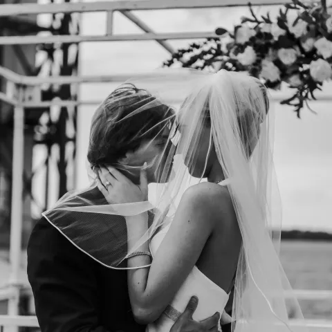 Bride and groom share a romantic kiss under a floral arch by the waterfront.