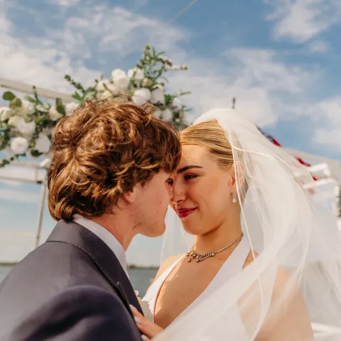 Bride and groom smiling, touching foreheads under a floral arch outside.