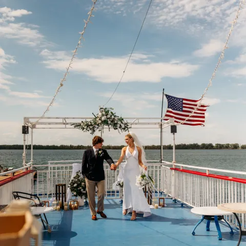 Couple walks on a boat deck under a cloudy sky with a U.S. flag in the background.