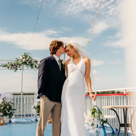 Bride and groom smiling on a deck with floral decor and blue sky.