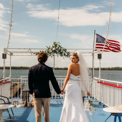 Bride and groom holding hands on a boat deck, American flag visible, blue sky background.